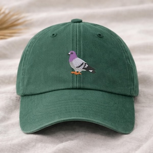 May include: A dark green baseball cap with a detailed embroidered image of a pigeon. The pigeon has a purple head, gray body, and orange feet. The cap has a curved brim and an adjustable strap.