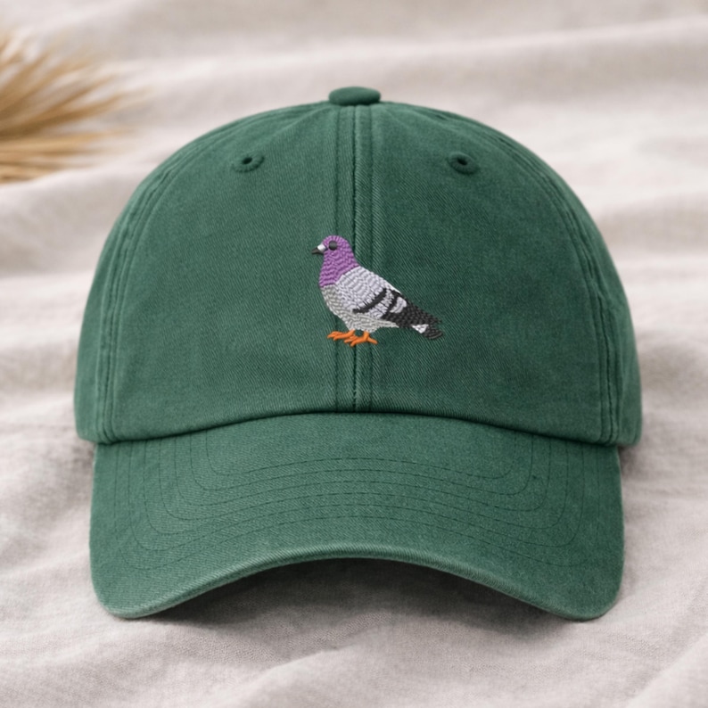 May include: A dark green baseball cap with a detailed embroidered image of a pigeon. The pigeon has a purple head, gray body, and orange feet. The cap has a curved brim and an adjustable strap.