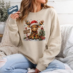 May include: A beige crewneck sweatshirt featuring a graphic of a cartoon reindeer wearing a red Santa hat, wrapped in colourful Christmas lights. The reindeer sits next to a North Pole sign, a decorated Christmas tree, and wrapped gift boxes.