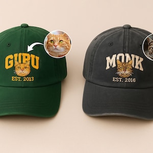 May include: Two baseball caps, one dark green and one charcoal grey. The green cap has "GURU" and "EST. 2013" embroidered with a cat face graphic. The grey cap has "MONK" and "EST. 2016" with a cat face graphic.