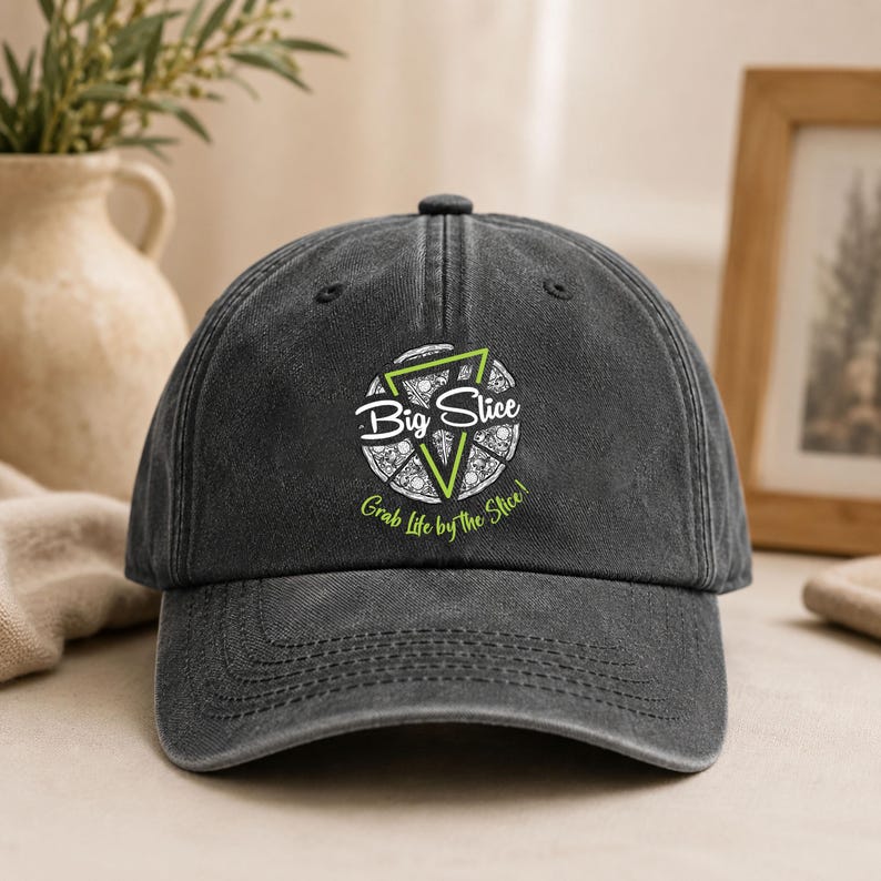 Puede incluir: Una gorra de b&eacute;isbol gris carb&oacute;n con un gr&aacute;fico en blanco y verde lima en la parte delantera. El dise&ntilde;o muestra una porci&oacute;n de pizza dentro de un tri&aacute;ngulo con el texto Big Slice y la frase Grab Life by the Slice! en blanco y verde.