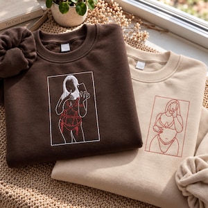 May include: A dark brown crewneck sweatshirt featuring a white and red embroidered outline of a person taking a mirror selfie in lingerie, paired with a matching brown scrunchie, and a beige sweatshirt with a red embroidered outline of a person.