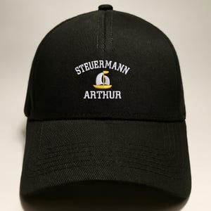 May include: Black baseball cap with the embroidered text "STEUERMANN ARTHUR" in white. A yellow and white sailboat graphic is centered below the word "STEUERMANN". The cap has a curved brim and a button on top.