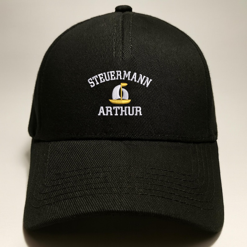 May include: Black baseball cap with the embroidered text "STEUERMANN ARTHUR" in white. A yellow and white sailboat graphic is centered below the word "STEUERMANN". The cap has a curved brim and a button on top.