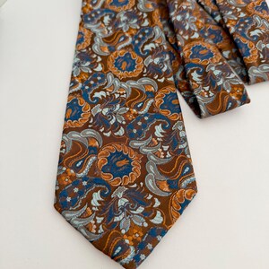 Könnte beinhalten: Eine Krawatte mit braunem Hintergrund und einem komplexen Webmuster in Hellblau, Dunkelblau und Orange. Das Design besteht aus sich wiederholenden floralen und paisleyartigen Motiven, die die gesamte Oberfläche des Stoffes bedecken.