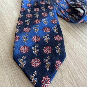 Puede incluir: Una corbata azul marino con un estampado repetido de flores rojas y hojas marrones. La corbata está hecha de una tela suave y es adecuada para ropa formal o de negocios. La corbata está colocada sobre una superficie de madera clara.