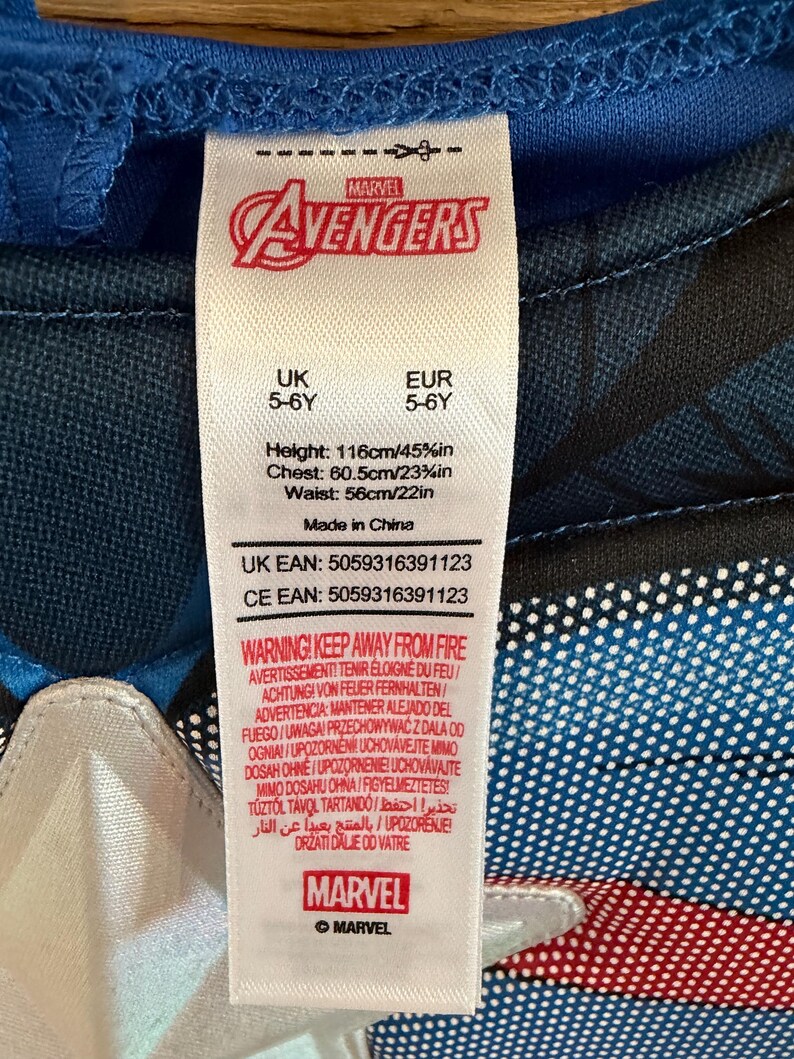 May include: A white clothing tag attached to a blue and black Marvel Avengers garment. The tag displays the Marvel logo, size 5-6Y, height 116cm/45.5in, chest 60.5cm/23.75in, waist 56cm/22in, Made in China, and a multi-language fire safety warning.