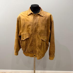 May include: A tan suede jacket with a collared neckline, two front pockets, and a button closure. The jacket is displayed on a black mannequin head and a metal stand against a gray background. The jacket has a ribbed hem and cuffs.