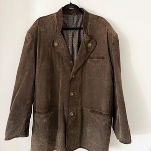 May include: A brown suede jacket with a notched collar and three buttons. The jacket has two front pockets and a patterned lining. The jacket is hanging on a black hanger against a white wall.