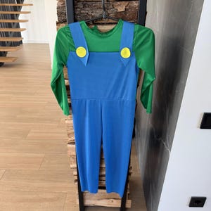May include: A long-sleeved jumpsuit featuring a bright green shirt top integrated with blue overalls. The blue overalls have two large, round, yellow buttons attached at the front where the straps meet the chest piece, creating a classic character look.