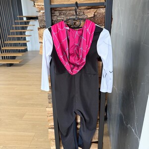 May include: A black and white Spider-Gwen costume with a pink hood featuring a web design. The costume has long white sleeves and a black jumpsuit. It is hanging on a black hanger.