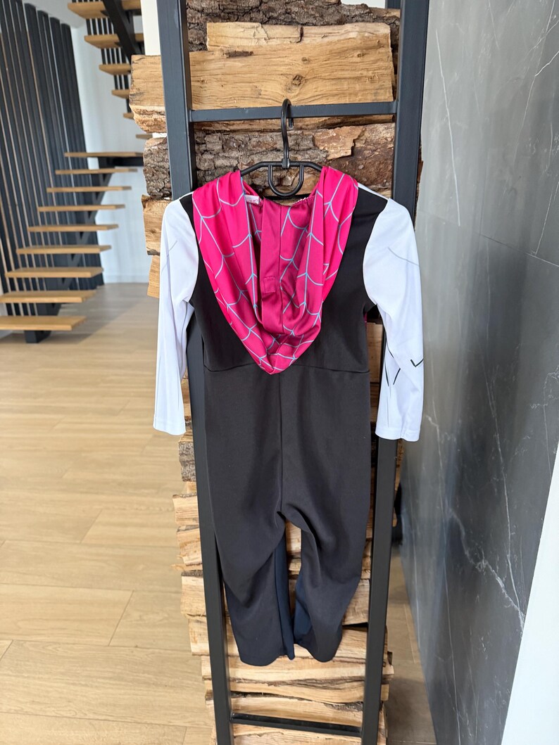 May include: A black and white Spider-Gwen costume with a pink hood featuring a web design. The costume has long white sleeves and a black jumpsuit. It is hanging on a black hanger.