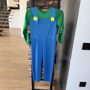 May include: A long-sleeved jumpsuit featuring a bright green shirt base with an attached blue overall overlay. The blue overalls include two prominent yellow circular buttons at the top of the straps, creating a distinct two-tone color block design.
