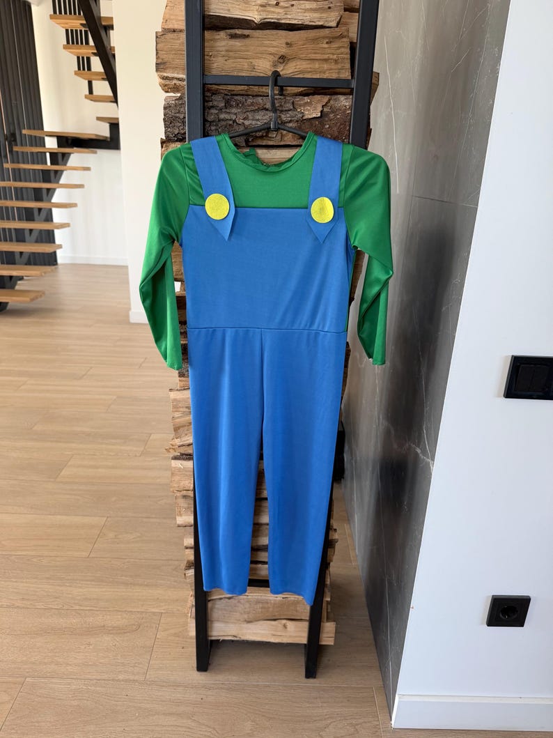 May include: A long-sleeved jumpsuit featuring a bright green shirt base with an attached blue overall overlay. The blue overalls include two prominent yellow circular buttons at the top of the straps, creating a distinct two-tone color block design.