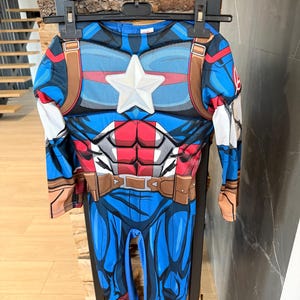 May include: A long-sleeved Captain America costume jumpsuit featuring a blue, red, and white color scheme. The chest displays a prominent white star emblem, printed muscle detailing, and brown belt graphics. The fabric has a smooth, printed finish.