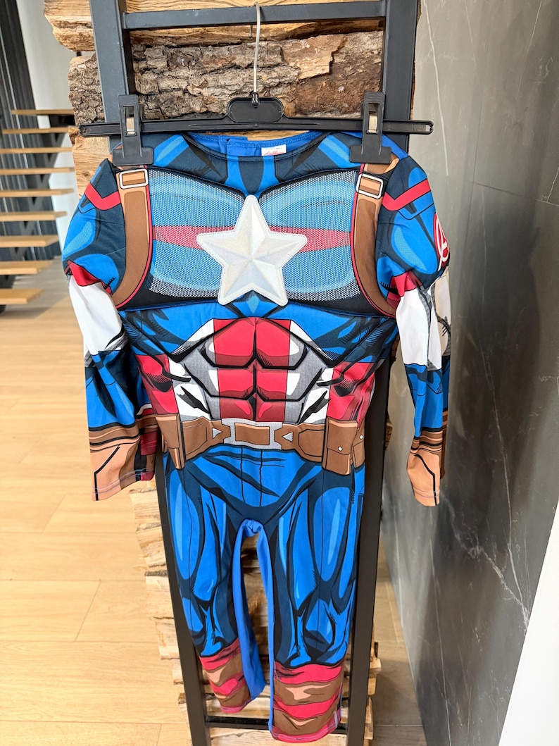 May include: A long-sleeved Captain America costume jumpsuit featuring a blue, red, and white color scheme. The chest displays a prominent white star emblem, printed muscle detailing, and brown belt graphics. The fabric has a smooth, printed finish.