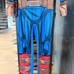 May include: A long-sleeved jumpsuit featuring a printed superhero costume design. The torso displays a red and grey chest plate graphic, while the legs are blue with brown boot-like details at the cuffs and a brown belt graphic at the waist.