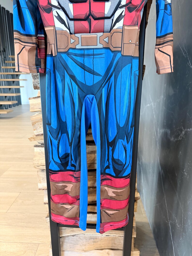 May include: A long-sleeved jumpsuit featuring a printed superhero costume design. The torso displays a red and grey chest plate graphic, while the legs are blue with brown boot-like details at the cuffs and a brown belt graphic at the waist.