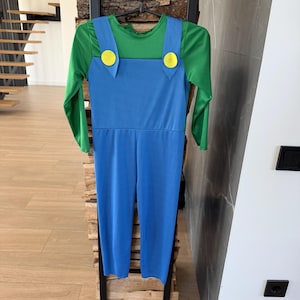 May include: A long-sleeved jumpsuit featuring a green shirt base with an attached blue overall overlay. The blue overalls include two prominent yellow circular buttons at the top of the straps, creating a distinct color-blocked appearance.