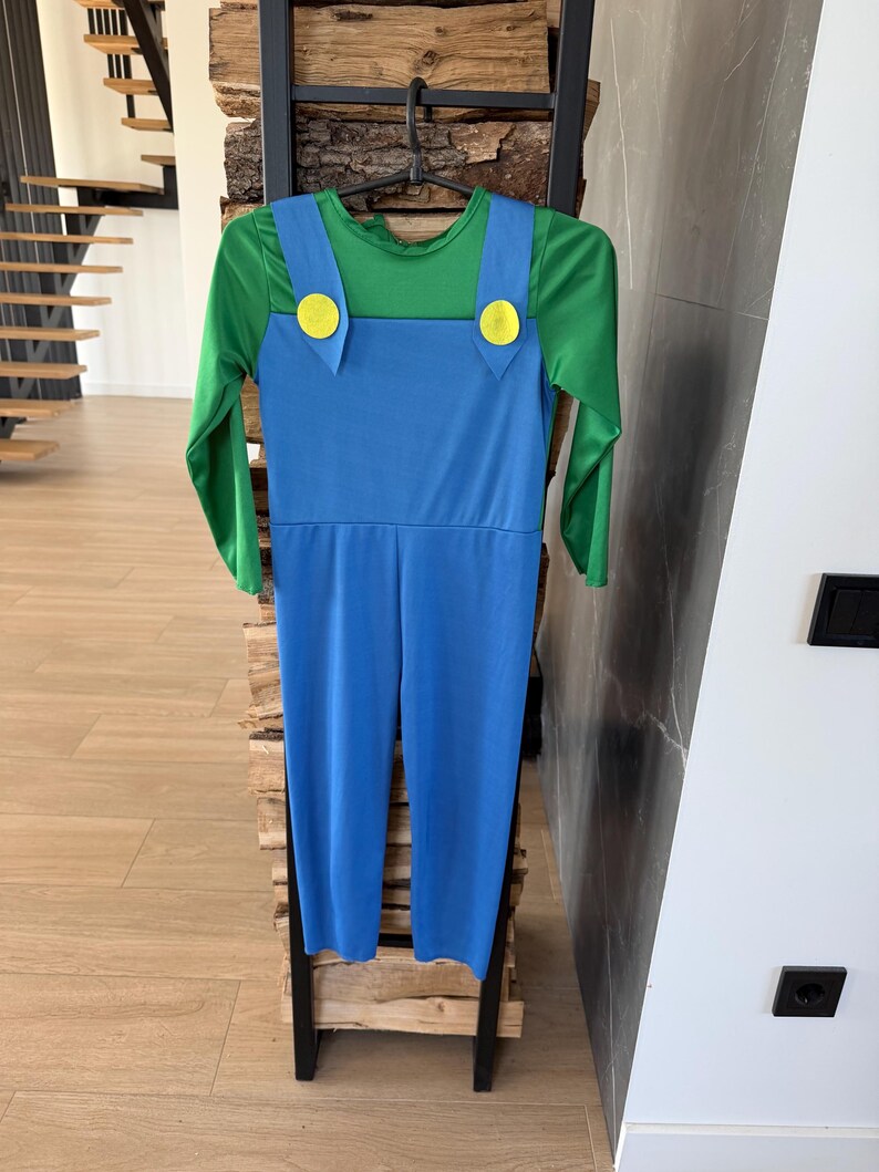May include: A long-sleeved jumpsuit featuring a green shirt base with an attached blue overall overlay. The blue overalls include two prominent yellow circular buttons at the top of the straps, creating a distinct color-blocked appearance.