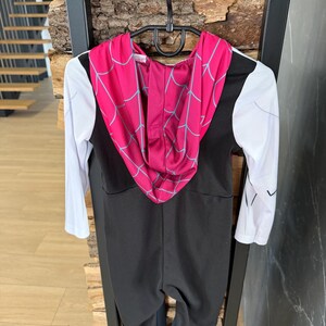 May include: A black and white Spider-Gwen costume for children. The costume has a black jumpsuit, white sleeves, and a pink hood with a blue spiderweb pattern. It is hanging on a black hanger.