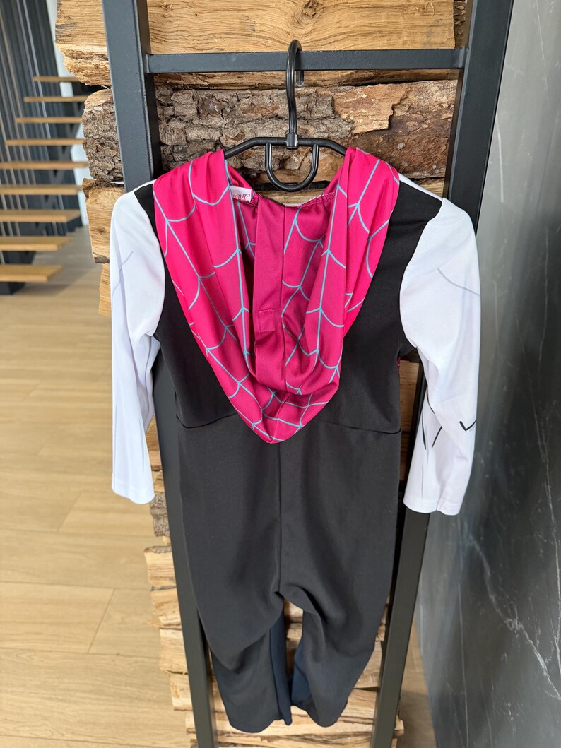May include: A black and white Spider-Gwen costume for children. The costume has a black jumpsuit, white sleeves, and a pink hood with a blue spiderweb pattern. It is hanging on a black hanger.