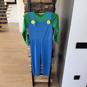 May include: A long-sleeved jumpsuit featuring a green shirt base with blue overalls attached. The blue overalls have two prominent yellow circular buttons at the top of the chest straps. The garment is displayed on a black plastic hanger.