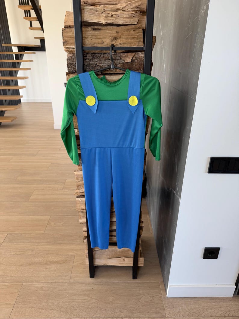 May include: A long-sleeved jumpsuit featuring a green shirt base with blue overalls attached. The blue overalls have two prominent yellow circular buttons at the top of the chest straps. The garment is displayed on a black plastic hanger.