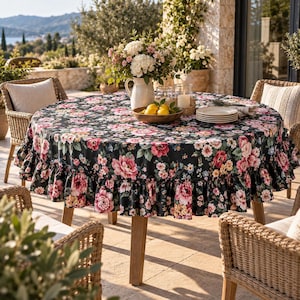 Op de afbeelding: Een rond tafelkleed met een zwarte achtergrond en een levendig bloemenpatroon van roze, rode en witte bloemen. Het tafelkleed heeft een geplooide rand en ligt over een ronde tafel, met daarop een witte kan, citroenen en borden.