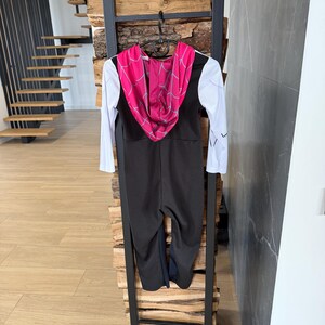 May include: A black and white jumpsuit with a pink hooded top featuring a spiderweb design. The costume is hanging on a black metal ladder-style display. The sleeves are white with black accents.