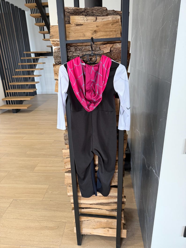 May include: A black and white jumpsuit with a pink hooded top featuring a spiderweb design. The costume is hanging on a black metal ladder-style display. The sleeves are white with black accents.