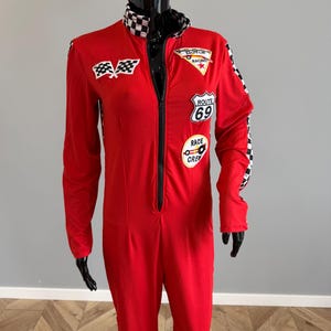 May include: A red long-sleeve jumpsuit featuring a black front zip, a chequered collar, and chequered side panels. The garment includes patches with the text Motor Racing, Route 69, and Race Crew, along with a chequered flag emblem on the chest.