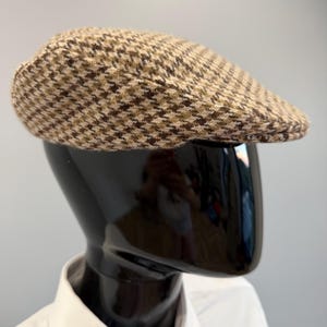 May include: A brown and tan houndstooth pattern flat cap. The cap is displayed on a black mannequin head, with a white collared shirt visible below. The cap's fabric has a textured appearance.