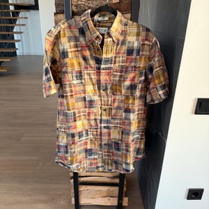 May include: A short-sleeved, button-down shirt with a patchwork plaid design in shades of yellow, red, blue, and brown. The shirt is hanging on a black hanger. The shirt is displayed on a small wooden stand.