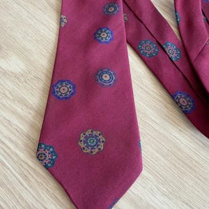 May include: A deep burgundy necktie featuring a repeating pattern of small, circular floral-like motifs. Each motif consists of a dark blue outer ring with teal, yellow, and orange accents arranged in a symmetrical, geometric design across the fabric.