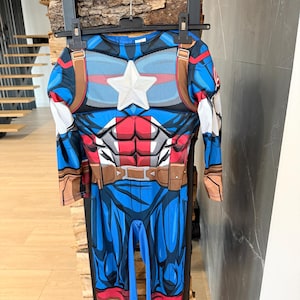 May include: A Captain America costume jumpsuit featuring a blue, red, and white color scheme with a prominent white star on the chest, printed muscle detailing, brown belt accents, and long sleeves, hanging on a black plastic clothes hanger.