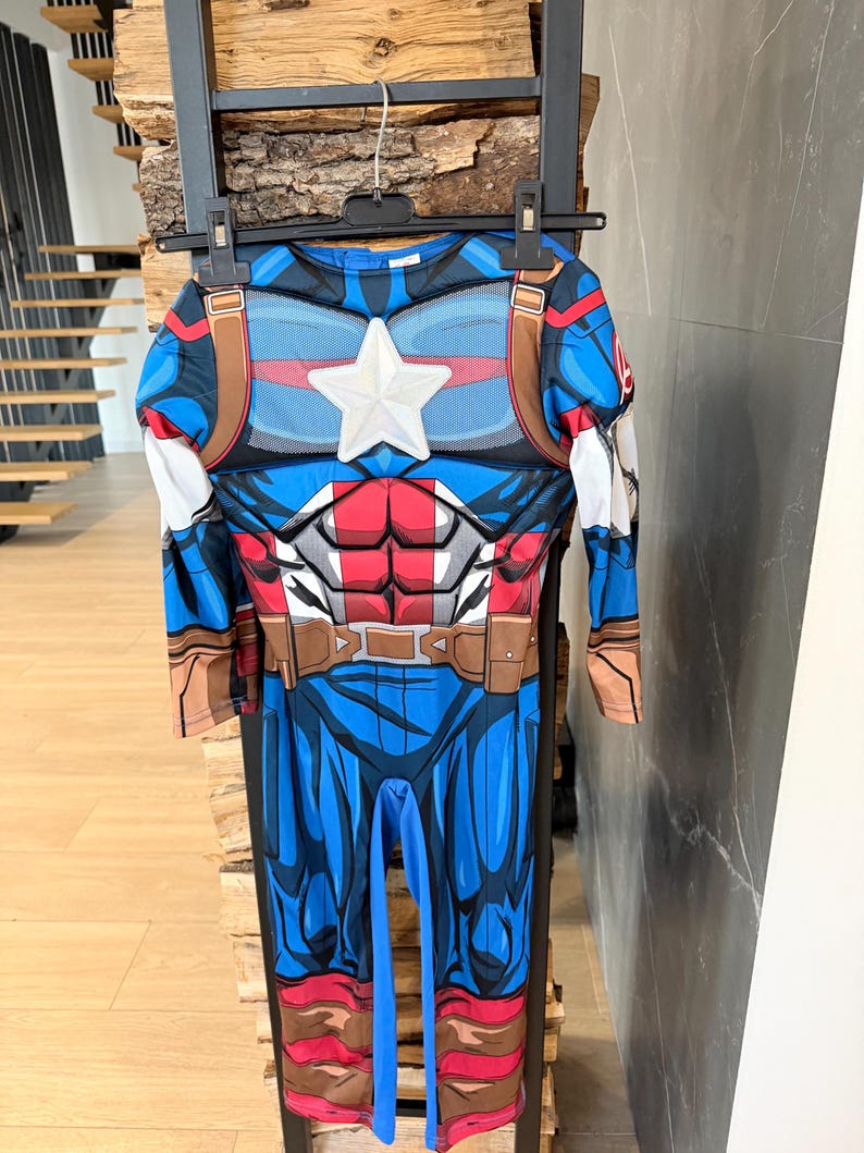 May include: A Captain America costume jumpsuit featuring a blue, red, and white color scheme with a prominent white star on the chest, printed muscle detailing, brown belt accents, and long sleeves, hanging on a black plastic clothes hanger.