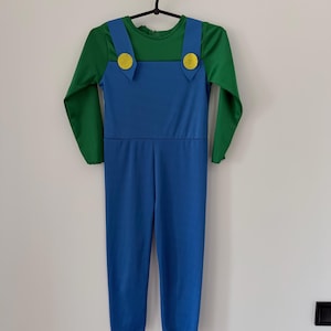 May include: A long-sleeved jumpsuit featuring a green shirt base with blue overalls attached. The blue overalls have two yellow circular buttons at the top of the straps. The garment is constructed from a smooth, solid-colored fabric.