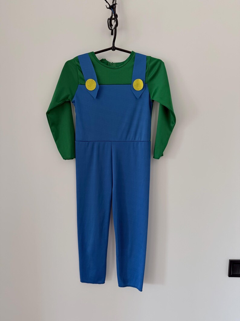 May include: A long-sleeved jumpsuit featuring a green shirt base with blue overalls attached. The blue overalls have two yellow circular buttons at the top of the straps. The garment is constructed from a smooth, solid-colored fabric.