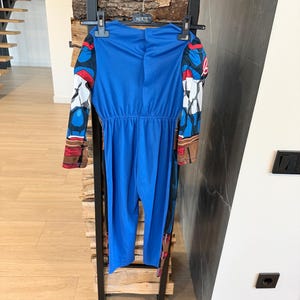 May include: A blue long-sleeved jumpsuit costume featuring printed graphic details on the sleeves, including red, white, and black patterns. The garment is suspended on a black plastic hanger with the word HERTE printed on the center hook area.
