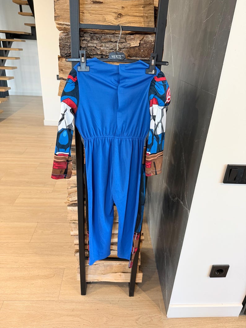 May include: A blue long-sleeved jumpsuit costume featuring printed graphic details on the sleeves, including red, white, and black patterns. The garment is suspended on a black plastic hanger with the word HERTE printed on the center hook area.