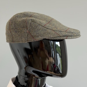 Può includere: Un berretto piatto in tweed verde oliva, con un motivo a quadri rossi e gialli. Presentato su una testa di manichino nera, ne evidenzia la forma classica e la trama. Un accessorio tradizionale e senza tempo.