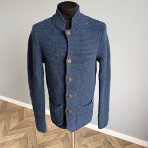 Può includere: Un cardigan blu navy in maglia con collo alto e bottoni in legno. Il cardigan ha due tasche frontali ed è esposto su un manichino. Il tessuto ha un aspetto strutturato.