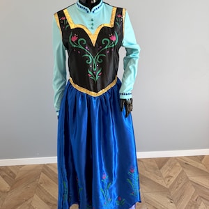 Pode incluir: Um vestido de fantasia com uma saia de cetim azul e um corpete preto com acabamentos dourados. O corpete tem um design floral com detalhes em rosa e verde. O vestido tem mangas compridas e uma parte superior azul clara.
