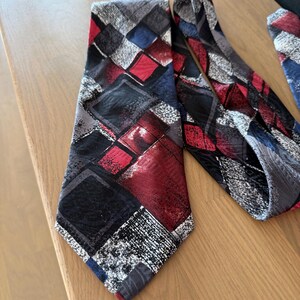 Peut inclure: Plusieurs cravates avec un motif géométrique. Les cravates présentent un motif répétitif de losanges rouges, noirs, bleus et gris. Les cravates sont faites d'une matière soyeuse et sont présentées sur une surface en bois.