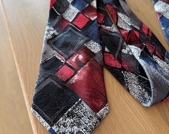 May include: A collection of neckties with a geometric pattern. The ties feature a repeating design of red, black, blue, and gray diamond shapes. The ties are made of a silky material and are displayed on a wooden surface.