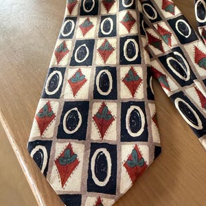 Op de afbeelding: Een stropdas met een patroon met een herhalend geometrisch ontwerp. Het patroon bevat zwarte ovalen, rode ruitvormen met groene accenten en zwarte vierkanten op een crèmekleurige achtergrond. De stropdas is gemaakt van een gladde stof en is geschikt voor formele of zakelijke kleding.