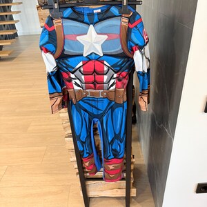 May include: A long-sleeved Captain America costume jumpsuit featuring a blue, red, and white color scheme. The chest displays a prominent white star, a printed muscular torso design, and a brown utility belt graphic. The Avengers logo is visible.