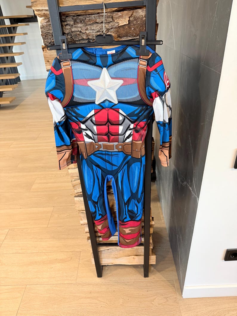 May include: A long-sleeved Captain America costume jumpsuit featuring a blue, red, and white color scheme. The chest displays a prominent white star, a printed muscular torso design, and a brown utility belt graphic. The Avengers logo is visible.