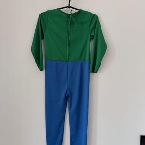 May include: A long-sleeved jumpsuit featuring a solid green top section and a solid blue bottom section. The garment has a round neckline with a tie closure at the back of the neck, long sleeves, and full-length pant legs, presented on a hanger.